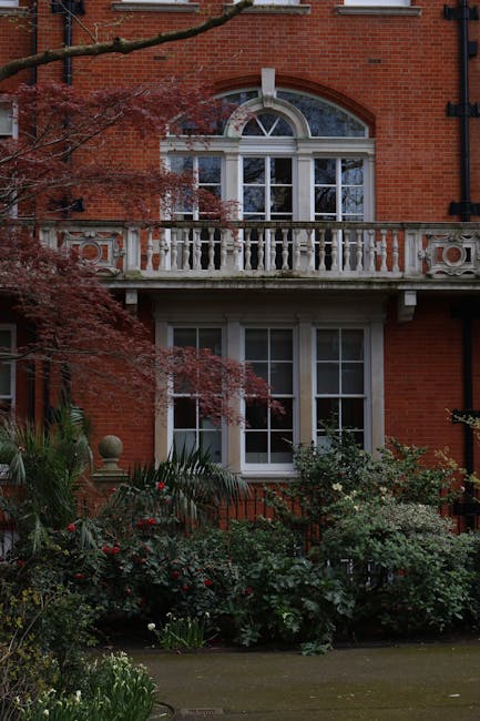 The image depicts the facade of a red brick Victorian-style building with a well-maintained garden in front. The garden features lush green shrubs, flowering plants, and small trees, creating a dense,